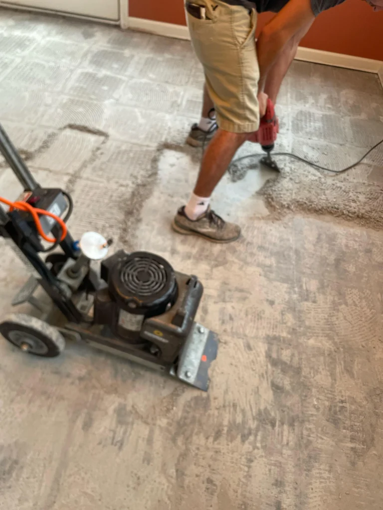 Worker using a concrete grinding machine to remove old adhesives and prepare the surface for new flooring in a residential setting.